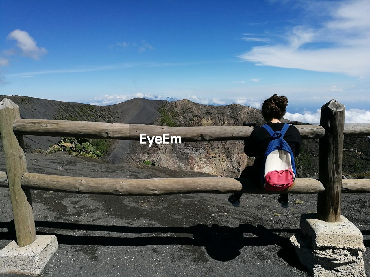 Rear view of young woman sitting on fence against mountains