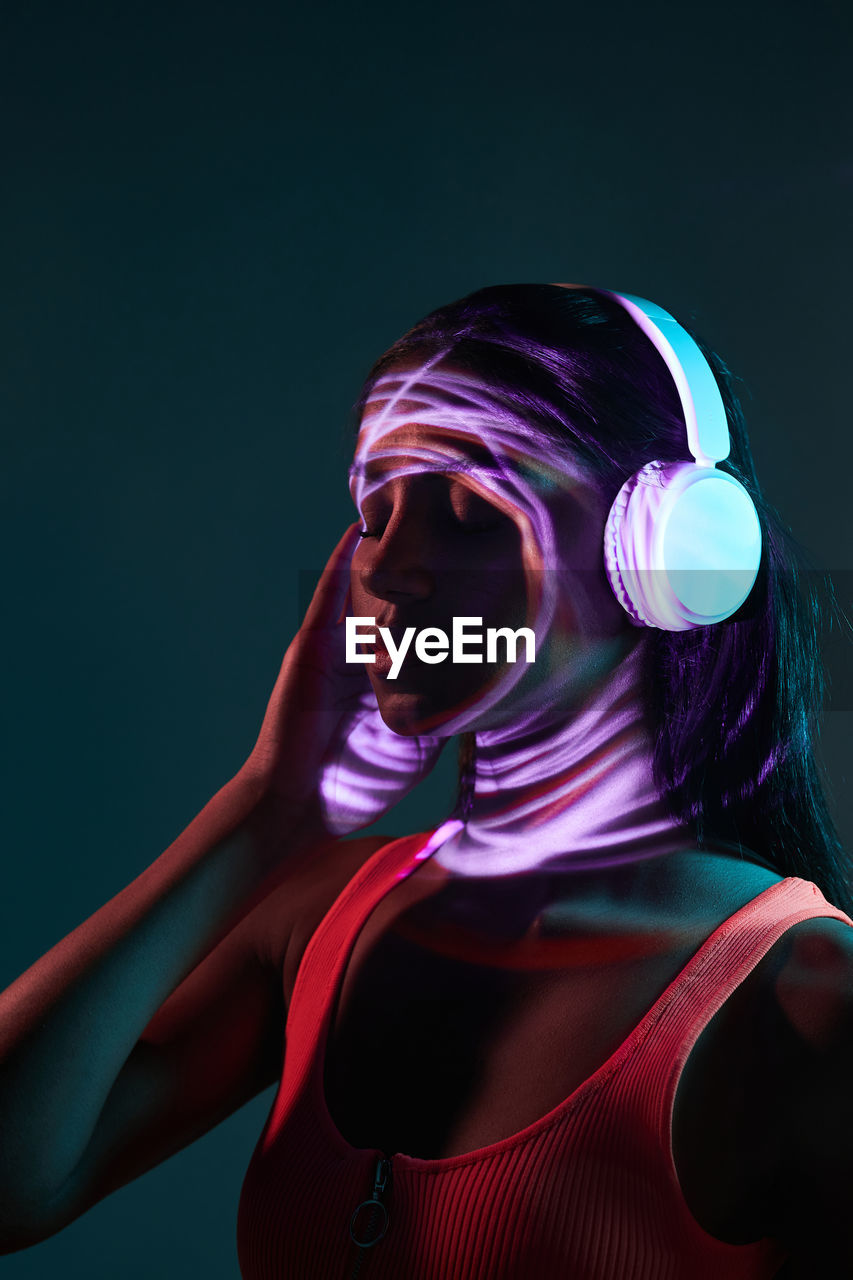 Charming brazilian female listening to music in wireless headphones while standing with eyes closed in obscure studio on dark background with shadow of light on face