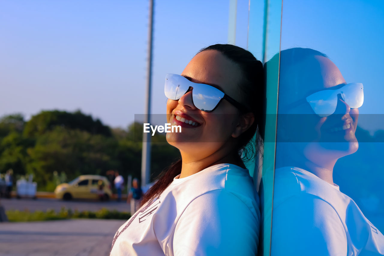 Smiling woman wearing sunglasses while leaning on wall