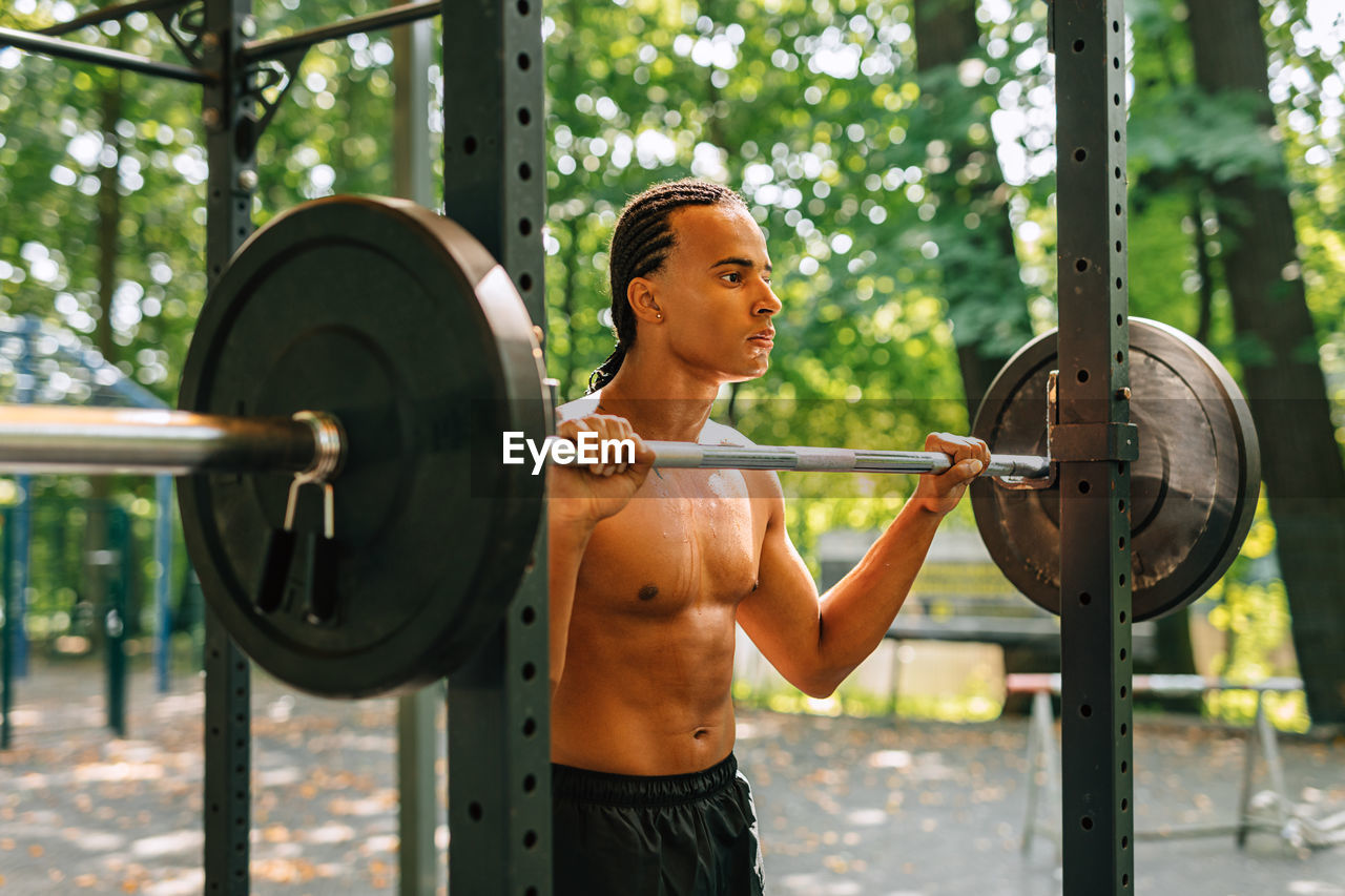 Shirtless man holding barbell while standing outdoors