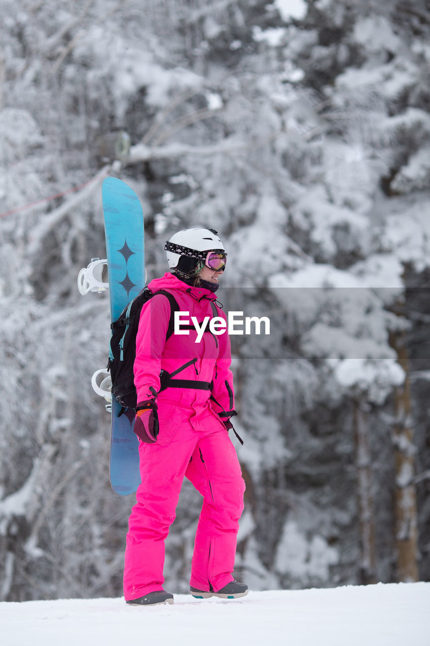 Woman in sportive overall with snowboard on backpack having fun in snowy day. winter escape concept.