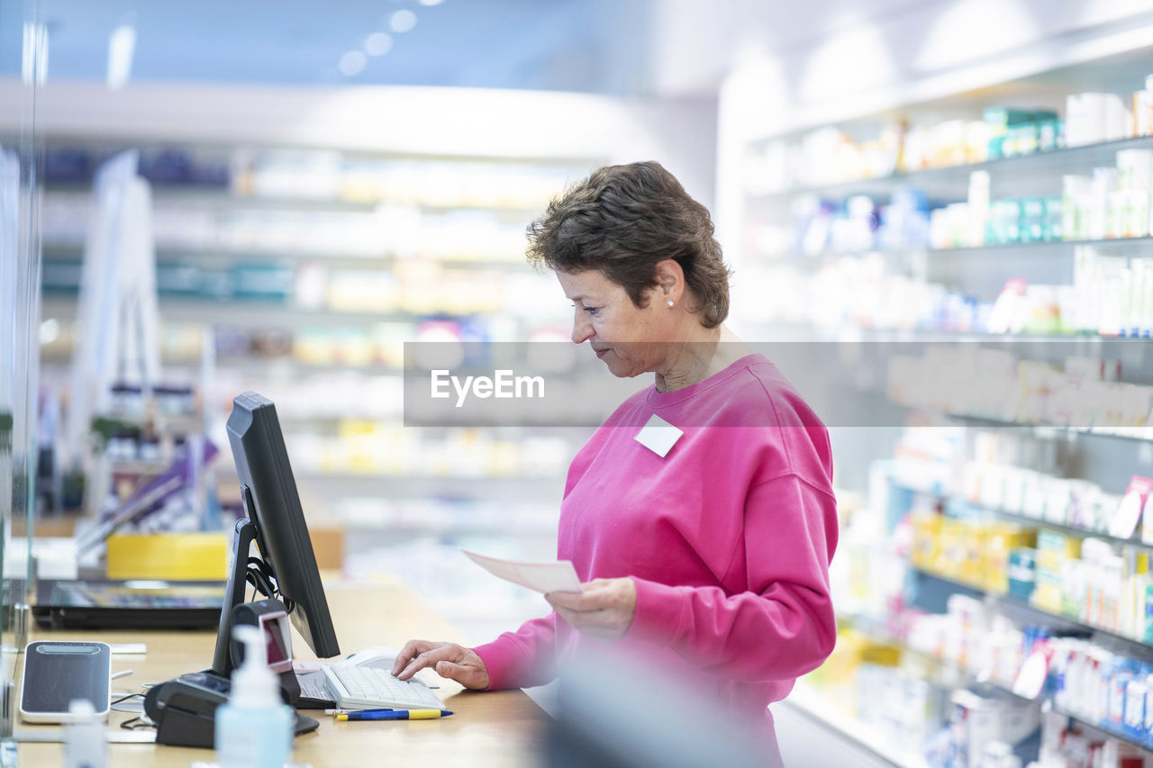 Smiling pharmacist with medical prescription using computer at desk