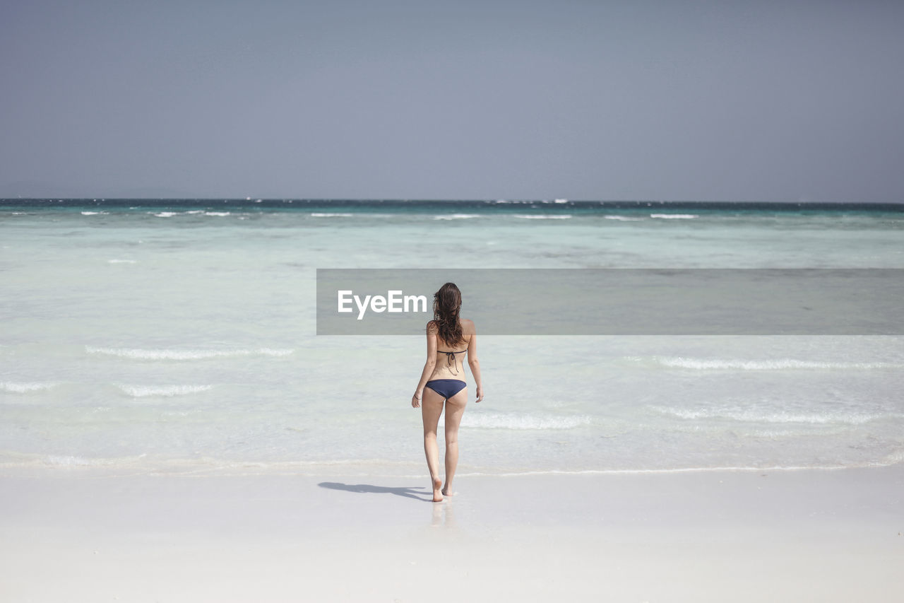 Full length rear view of woman in biking walking towards sea against clear sky