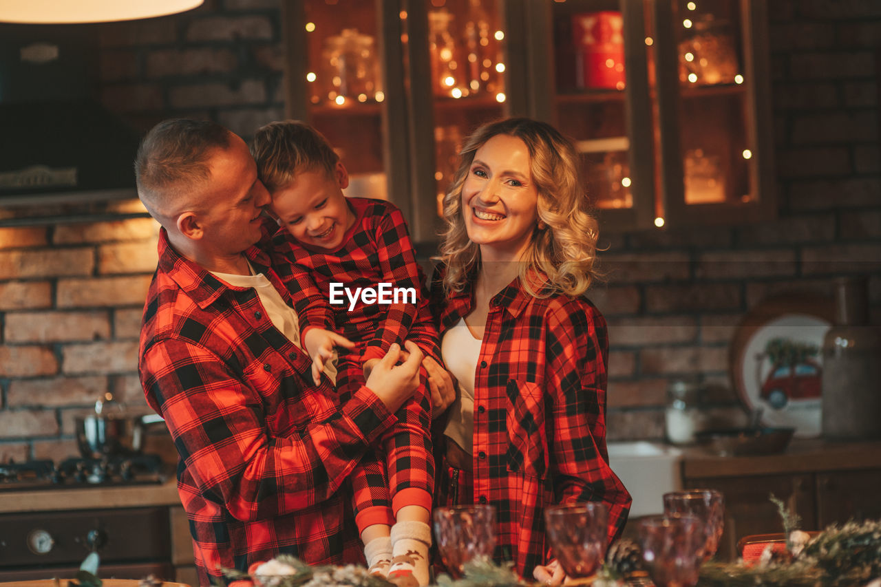 Smile active parents with small son in red checkered sleepwears waiting santa indoor.
