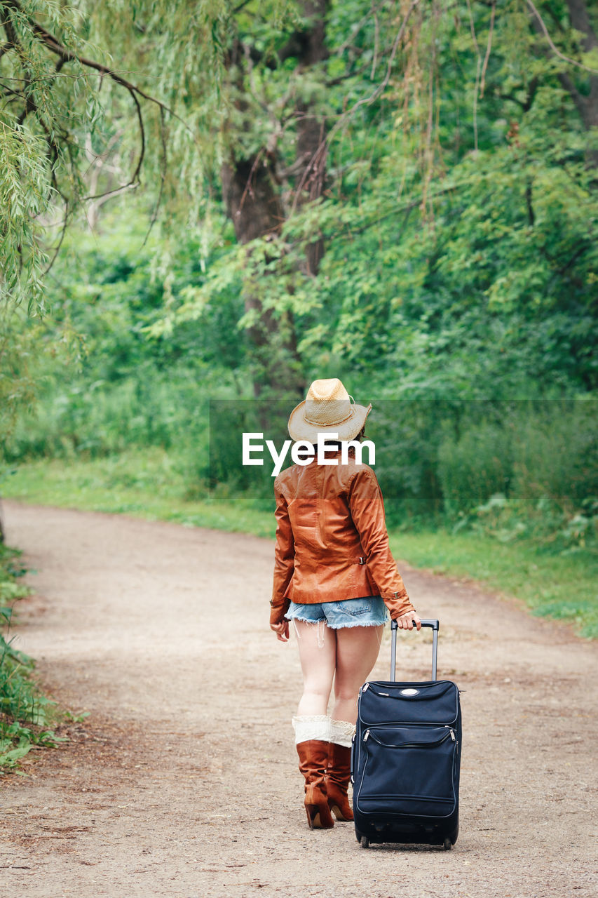 Rear view of woman pulling suitcase on dirt road against trees