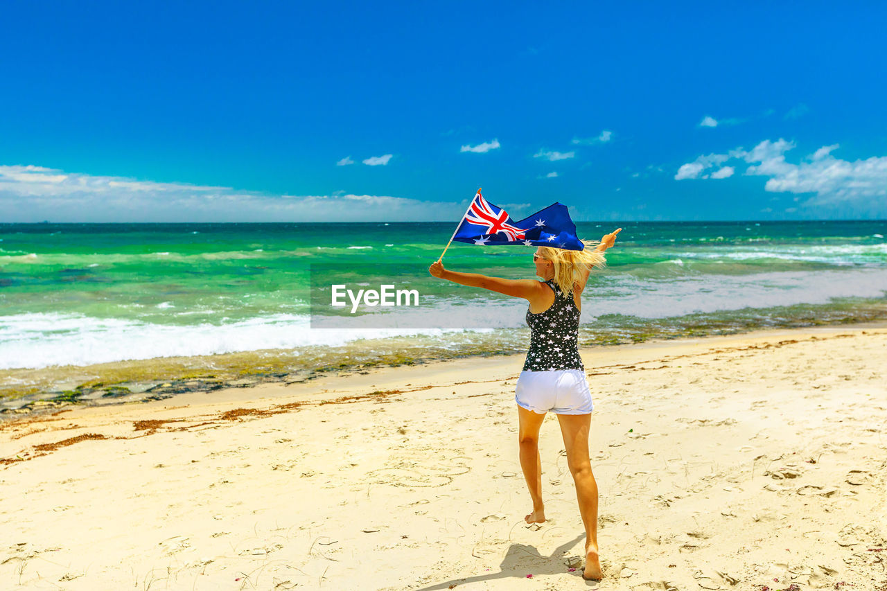 Rear view full length of woman holding australian flag running at beach