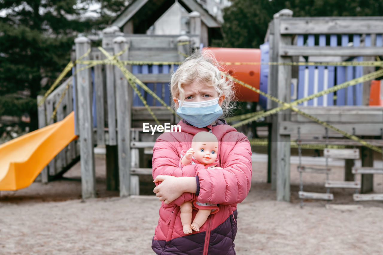 Sad caucasian girl in face mask with baby toy on closed playground outdoor. 