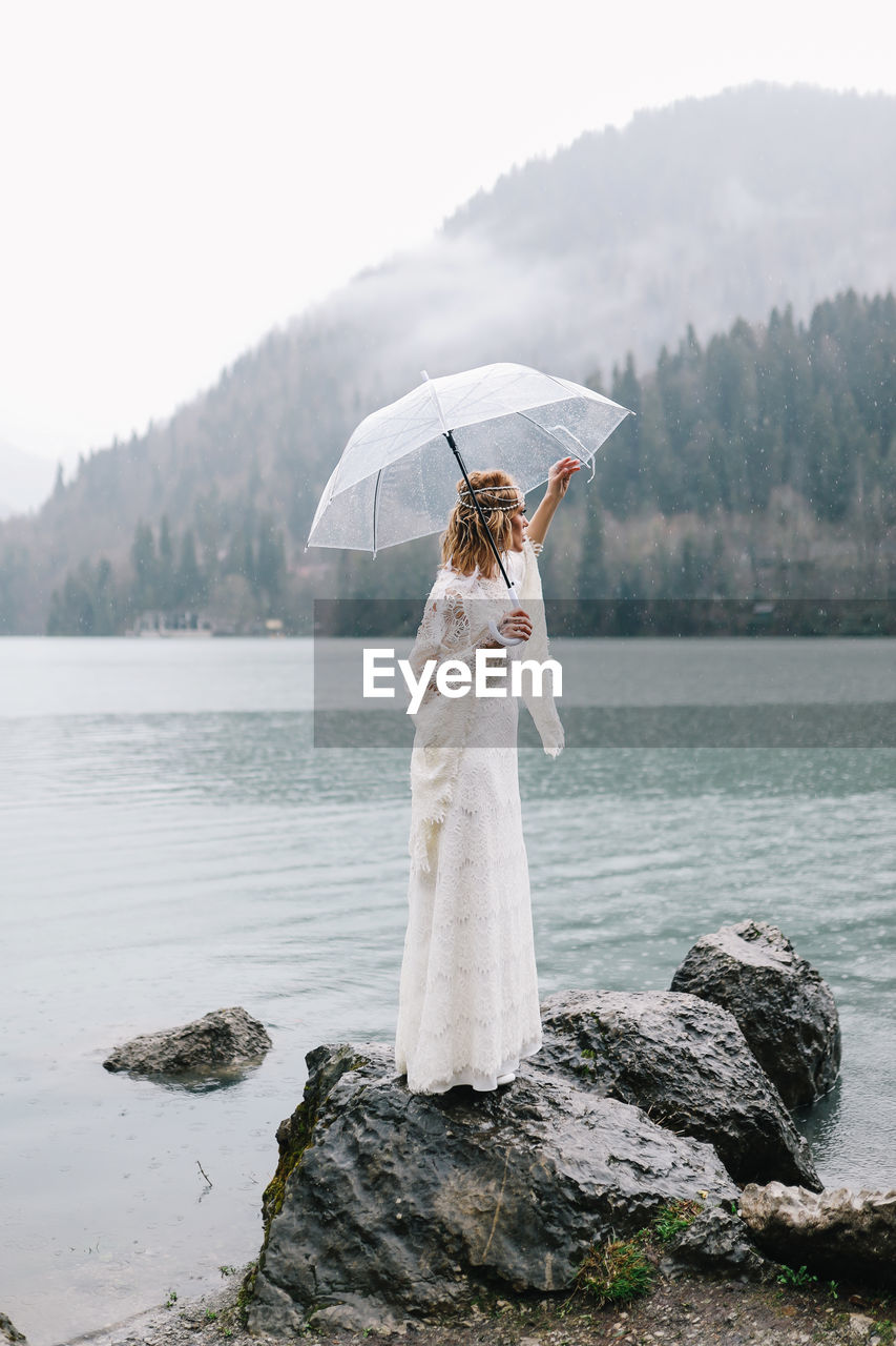 A beautiful young woman bride in a wedding lace dress stands in the middle of a lake and mountains