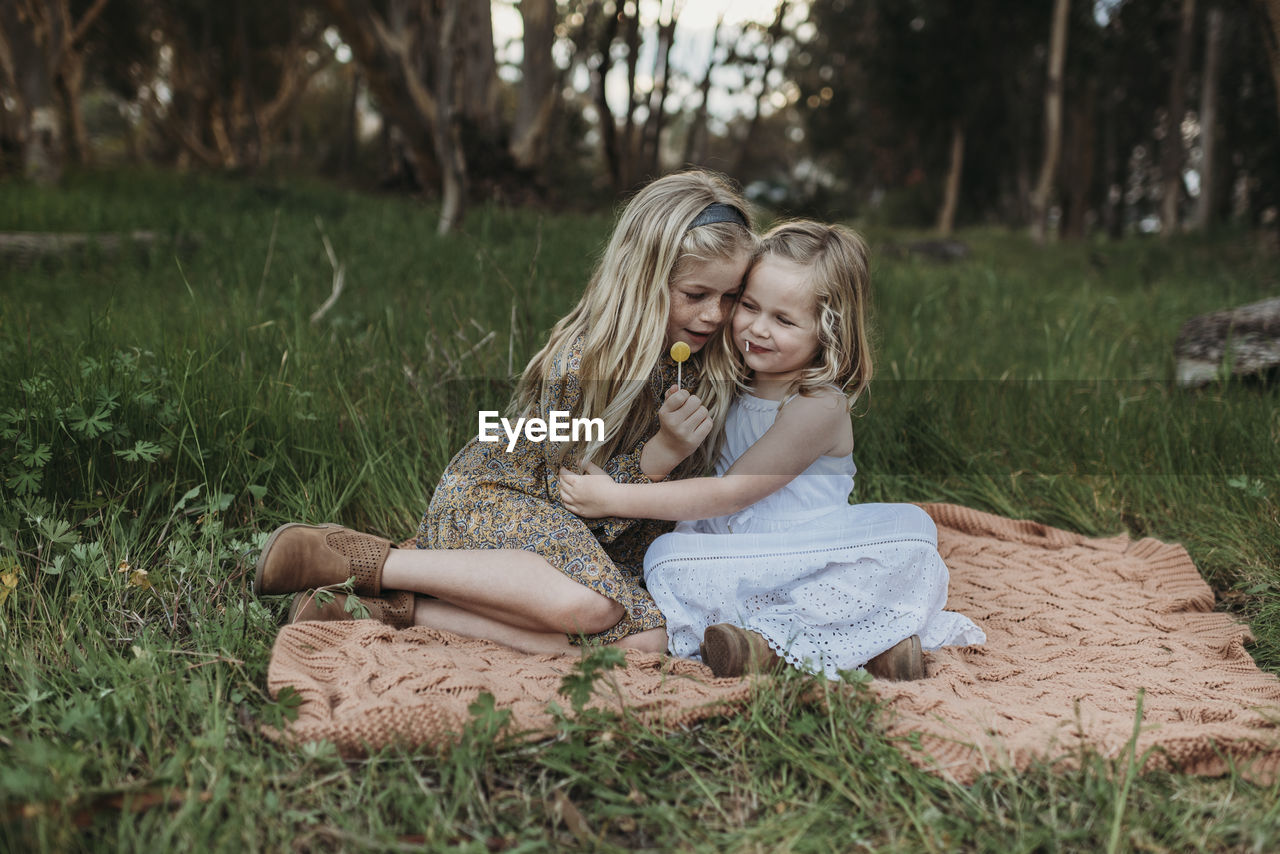 Two blond sisters sitting on a blanket in a field with lollipops