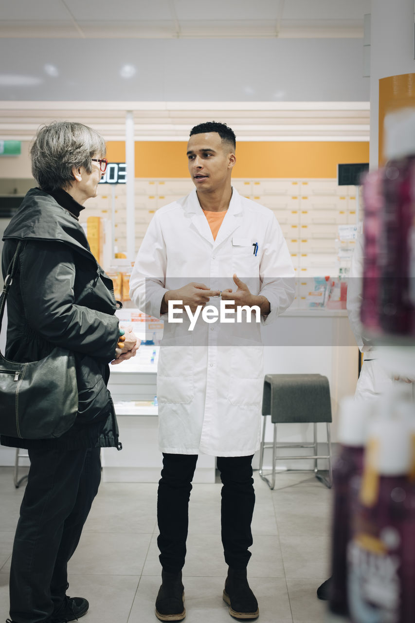 Full length of male pharmacist talking with senior female customer in medical store