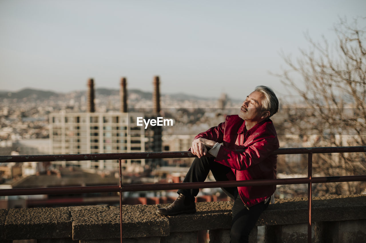 Relaxed mature man sitting on retaining wall with city in background