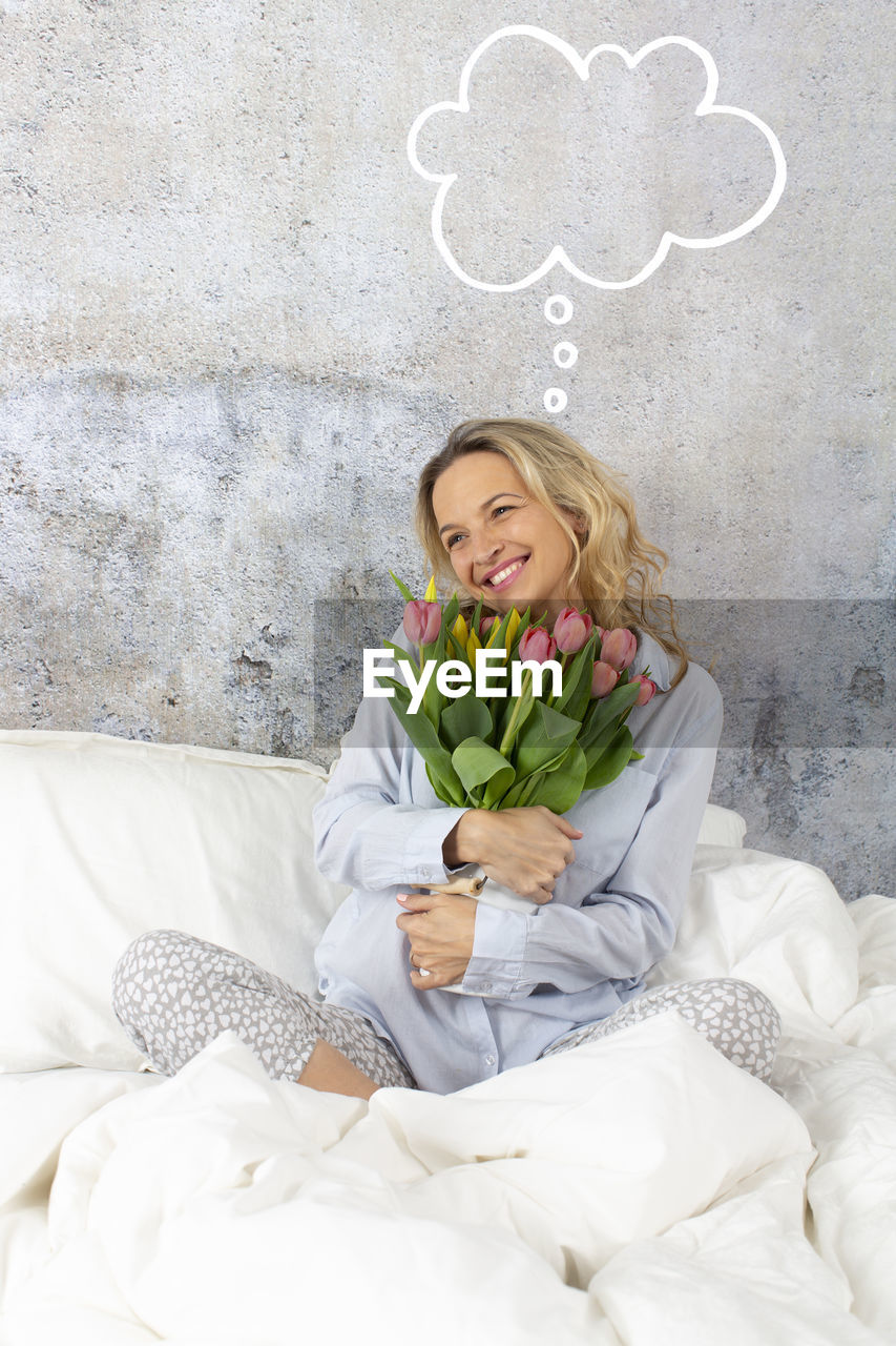 Smiling woman holding bouquet with speech bubble symbol on bed at home