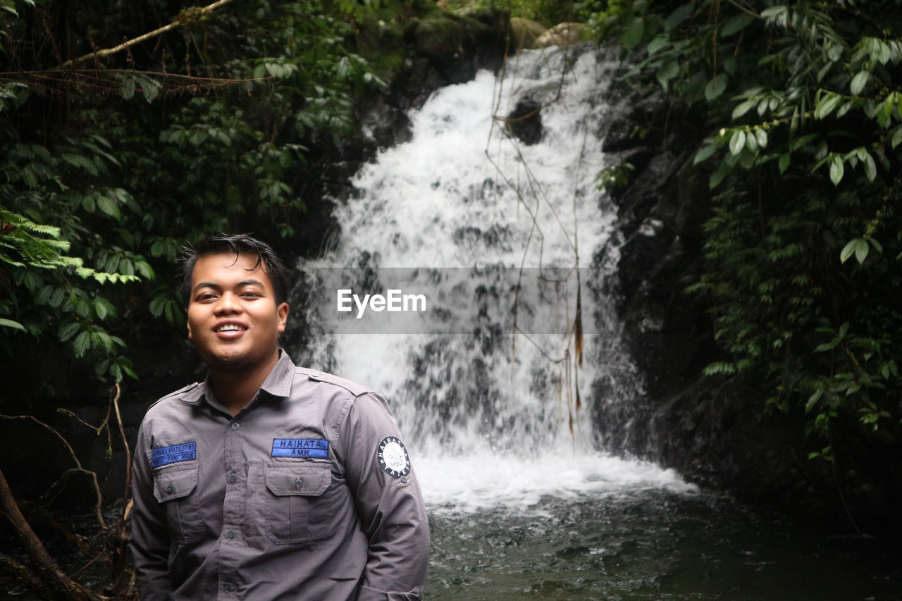 Portrait of smiling young man standing against waterfall in forest