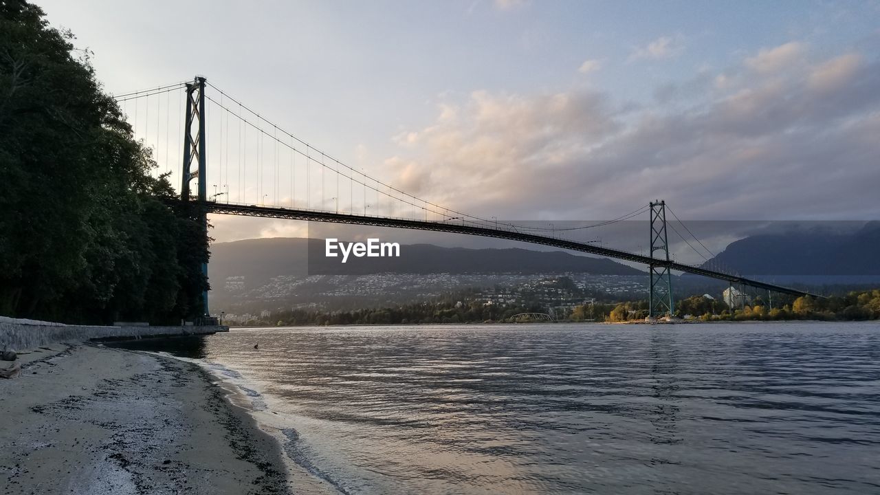 VIEW OF SUSPENSION BRIDGE OVER RIVER AGAINST CLOUDY SKY