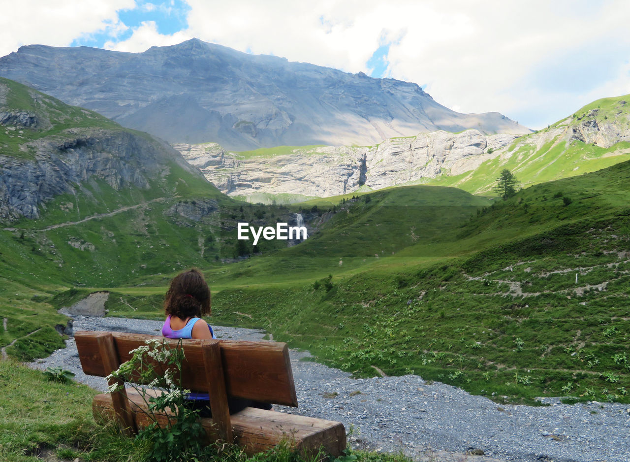 Woman sitting on wooden bench, looking at scenic view of mountains in summer 