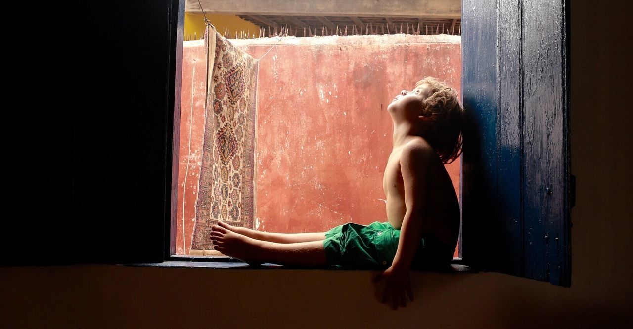 Full length of shirtless boy sitting on window sill at home