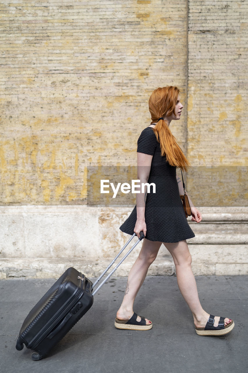 Young woman with suitcase walking next to stone wall