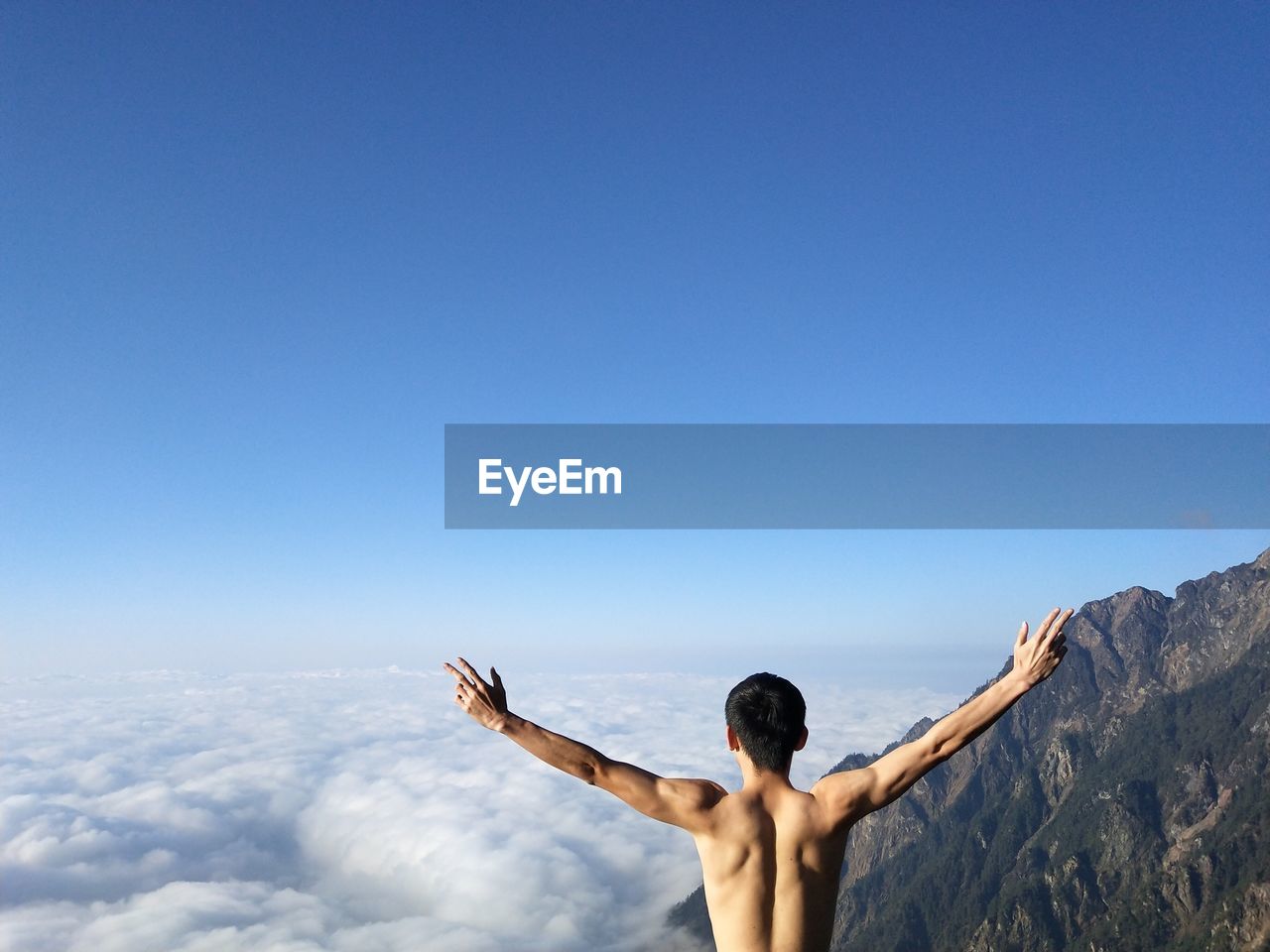 Rear view of shirtless man with arms outstretched standing against blue sky