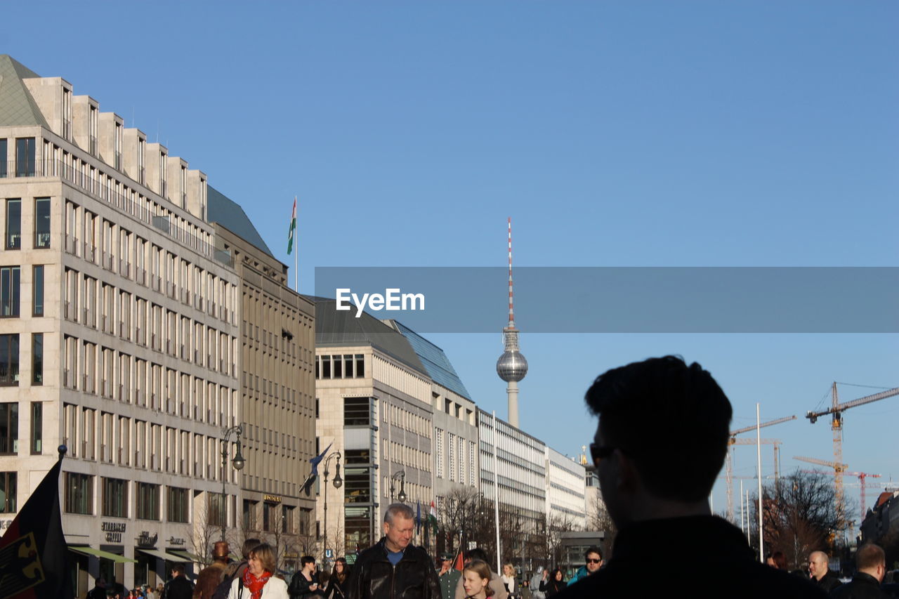 People walking on street by buildings and fernsehturm against blue sky