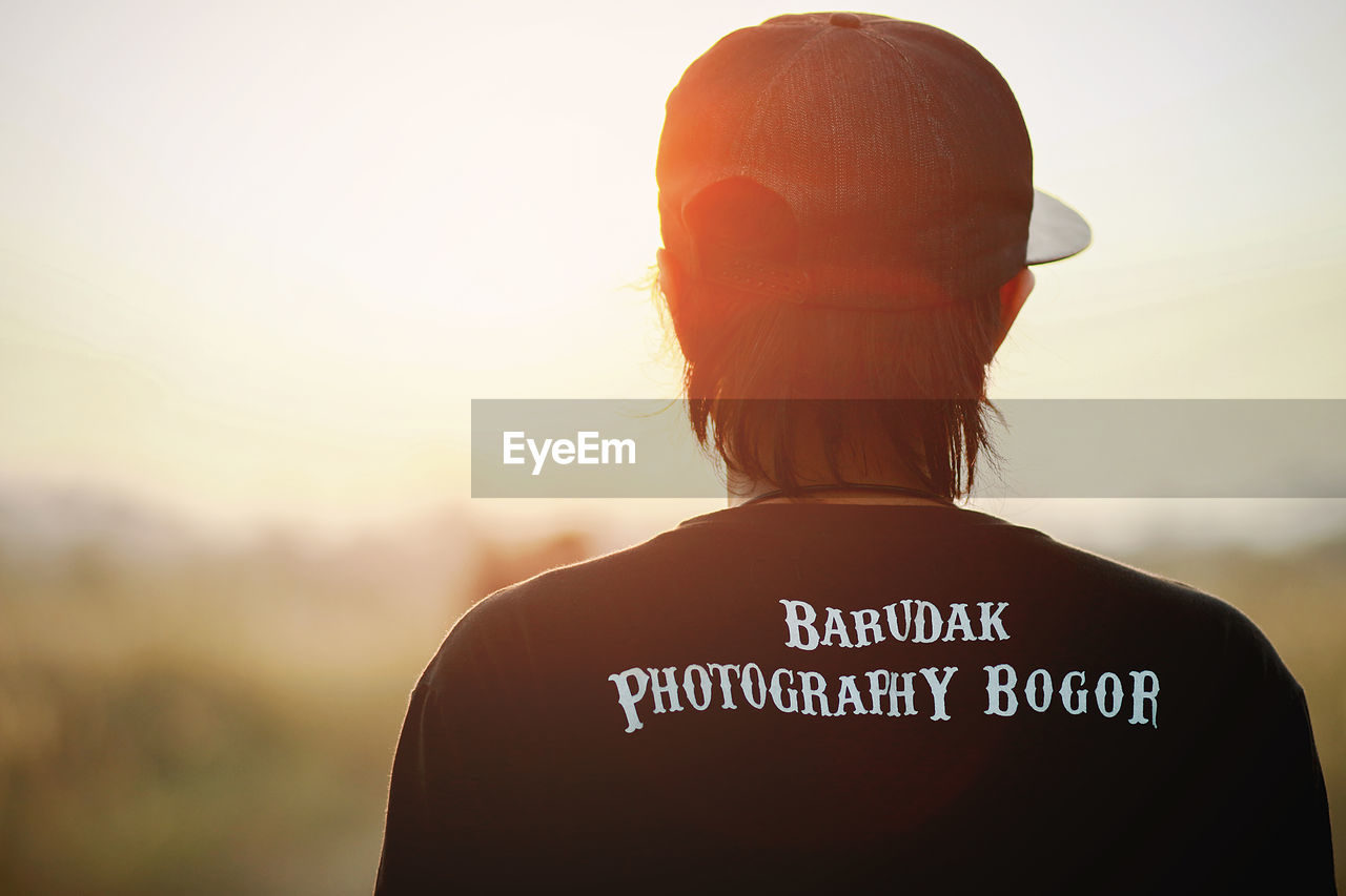 REAR VIEW OF WOMAN STANDING AGAINST SKY AT SUNSET