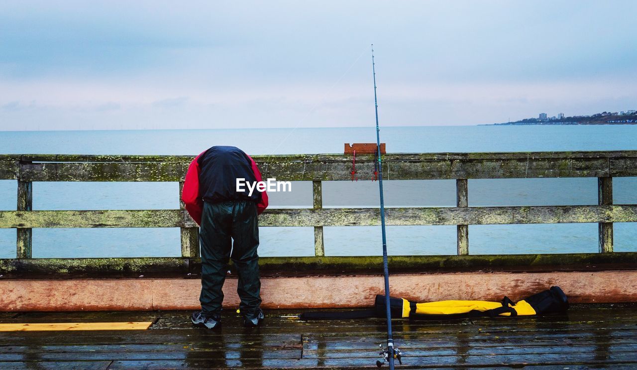 Rear view of man bending on pier over sea against sky