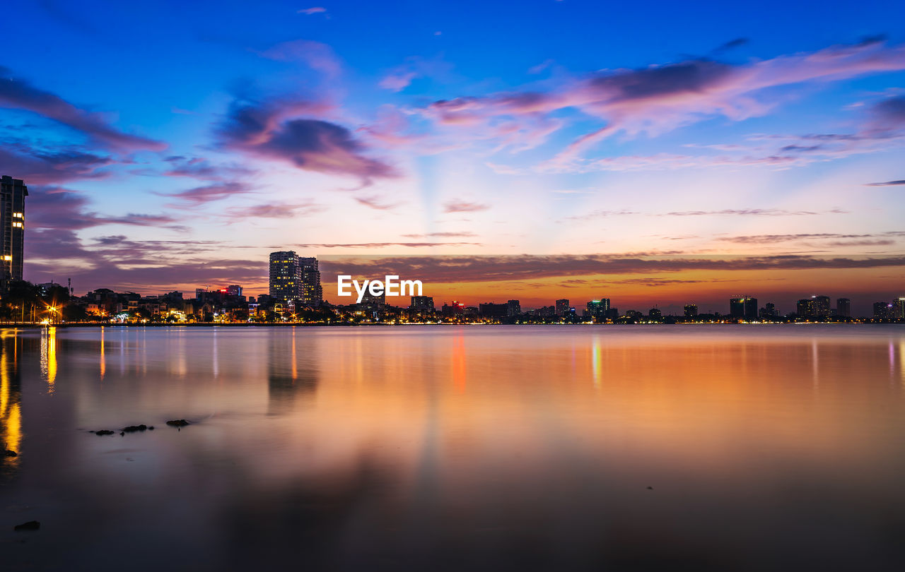 Scenic view of lake by buildings against sky during sunset