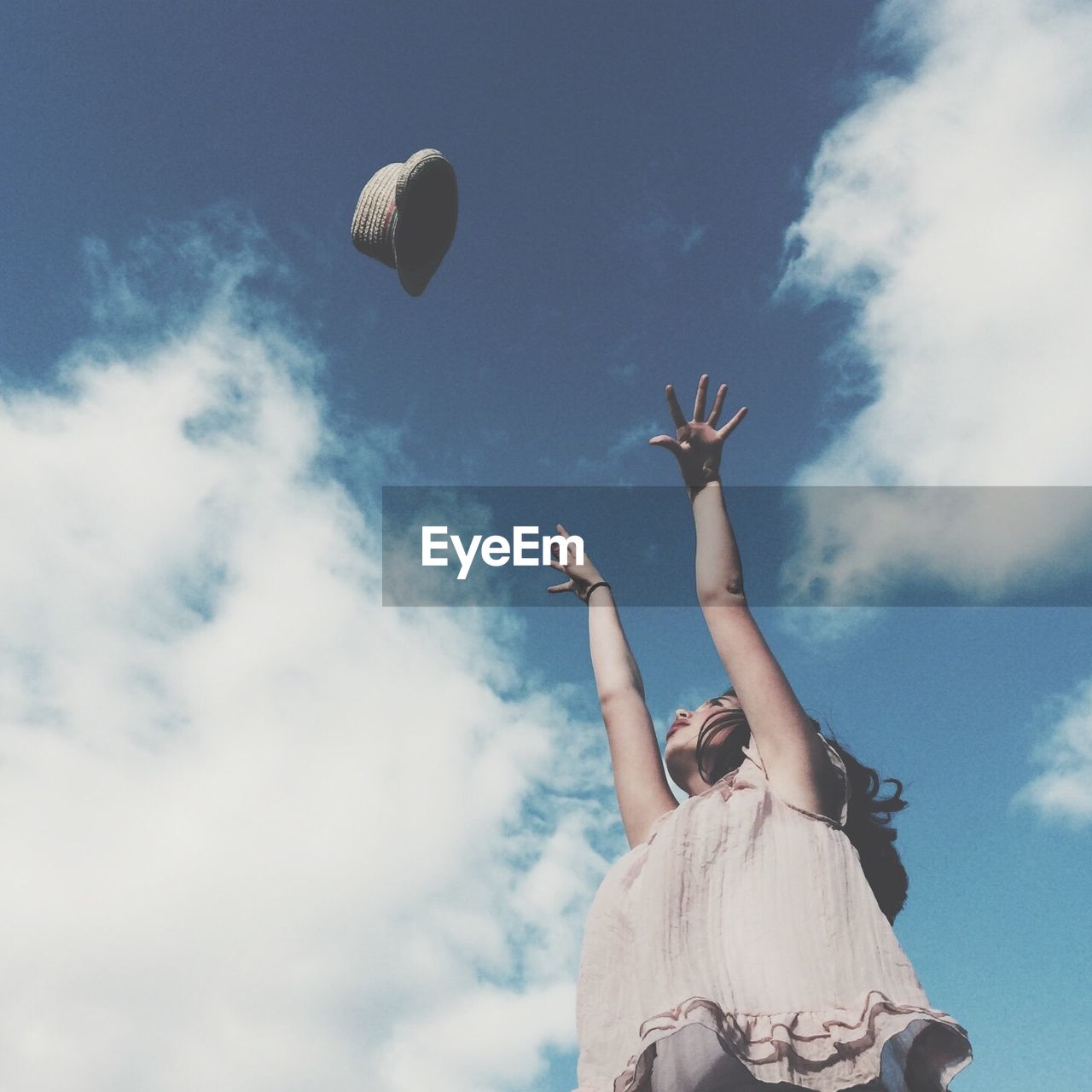 Low angle view of woman throwing hat in mid-air against cloudy sky