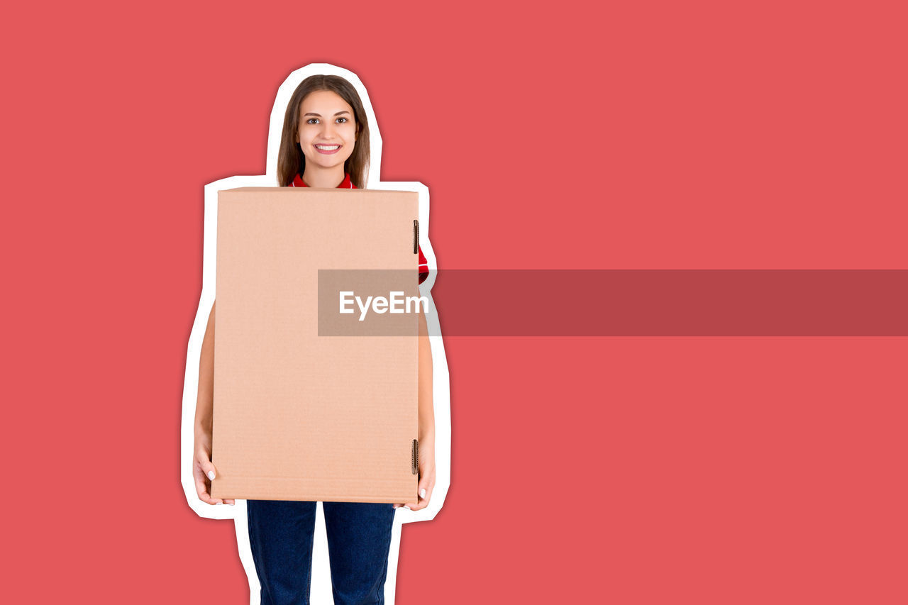 Portrait of smiling young woman with cardboard boxes standing against red background