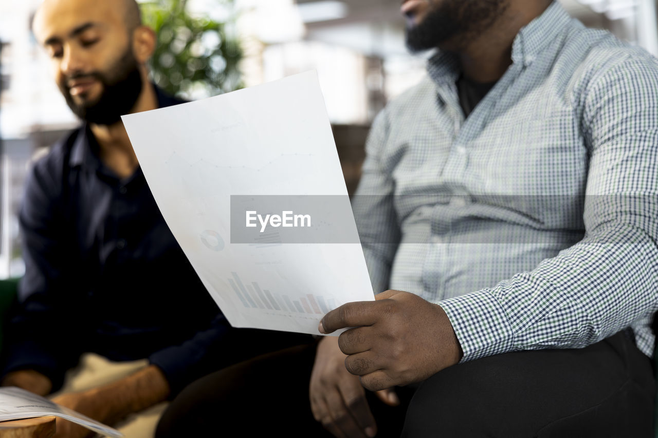 adult, men, sitting, business, paper, two people, young adult, conversation, communication, indoors, person, businessman, business finance and industry, document, occupation, activity, planning, cooperation, holding, office, looking, working, teamwork, meeting, creativity, professional occupation, education, casual clothing, clothing, facial hair, reading, table, beard, businesswear