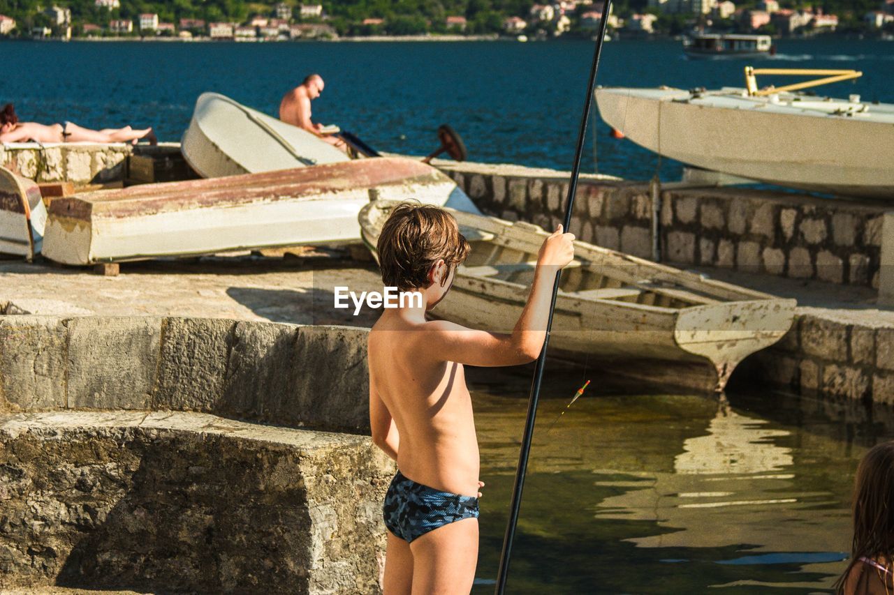 REAR VIEW OF SHIRTLESS MAN STANDING AT HARBOR