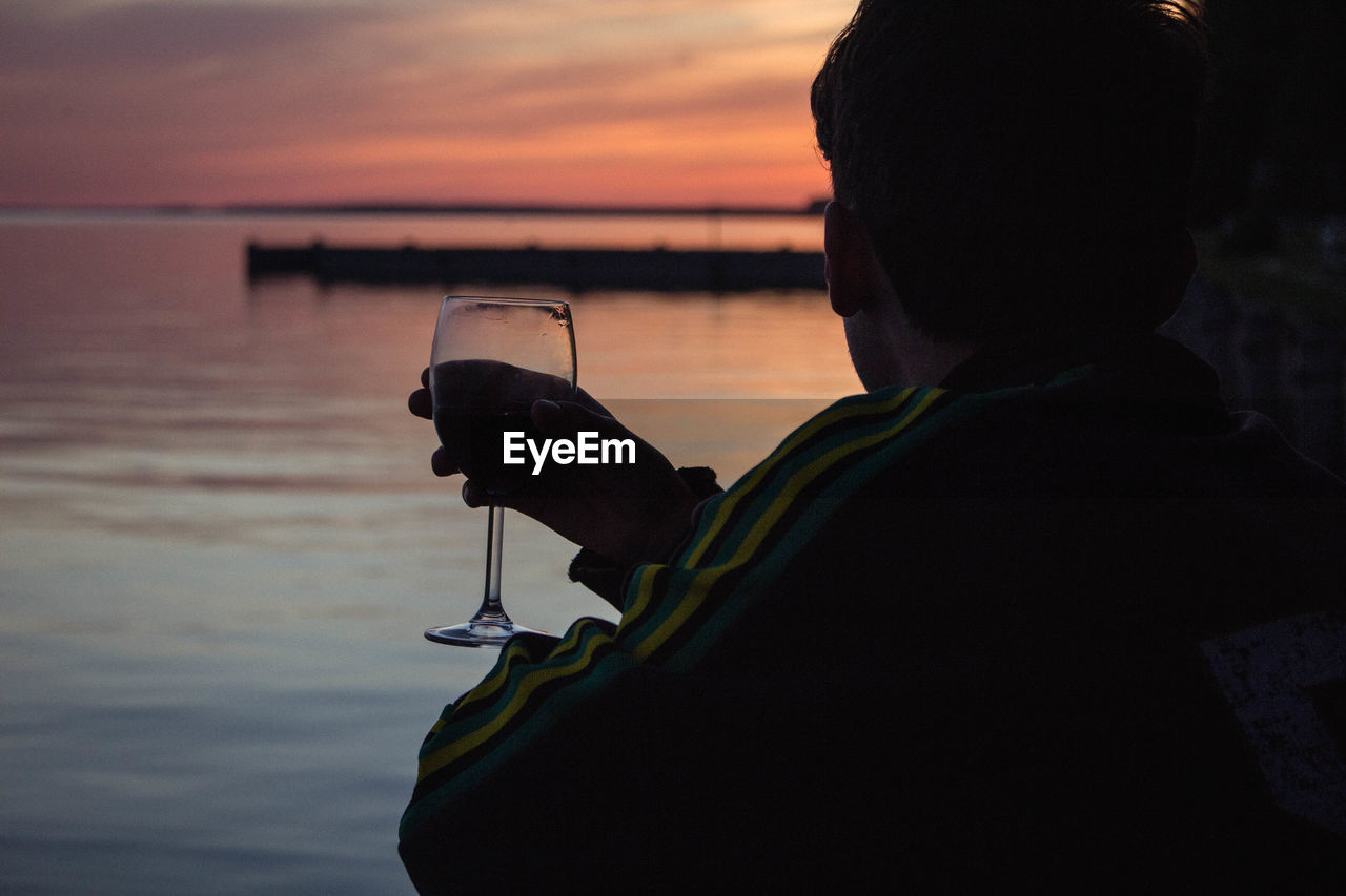 Man drinking wine by sea against sky during sunset