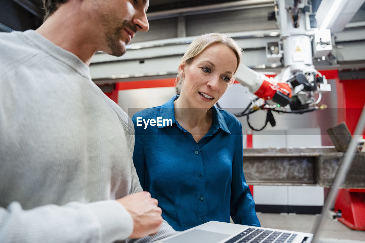 Mature engineer looking at laptop held by colleague in robot factory