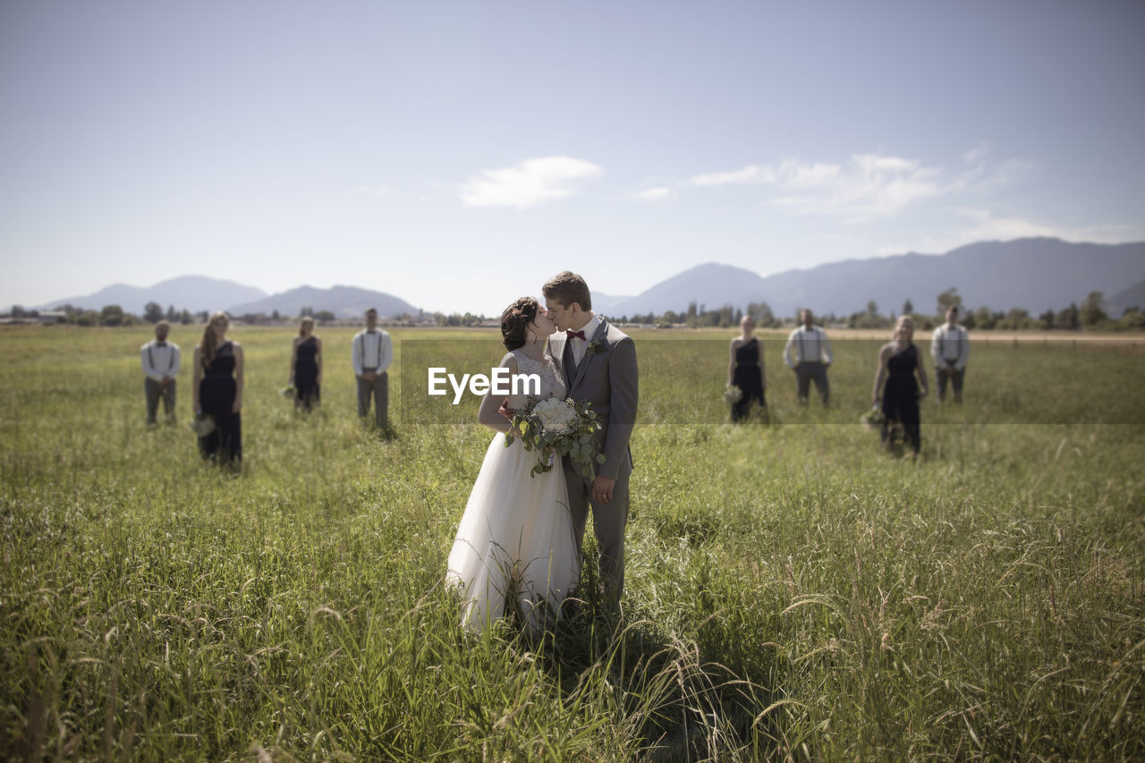 Newlywed couple kissing while standing at grassy field with groomsmen and bridesmaids in background during sunny day