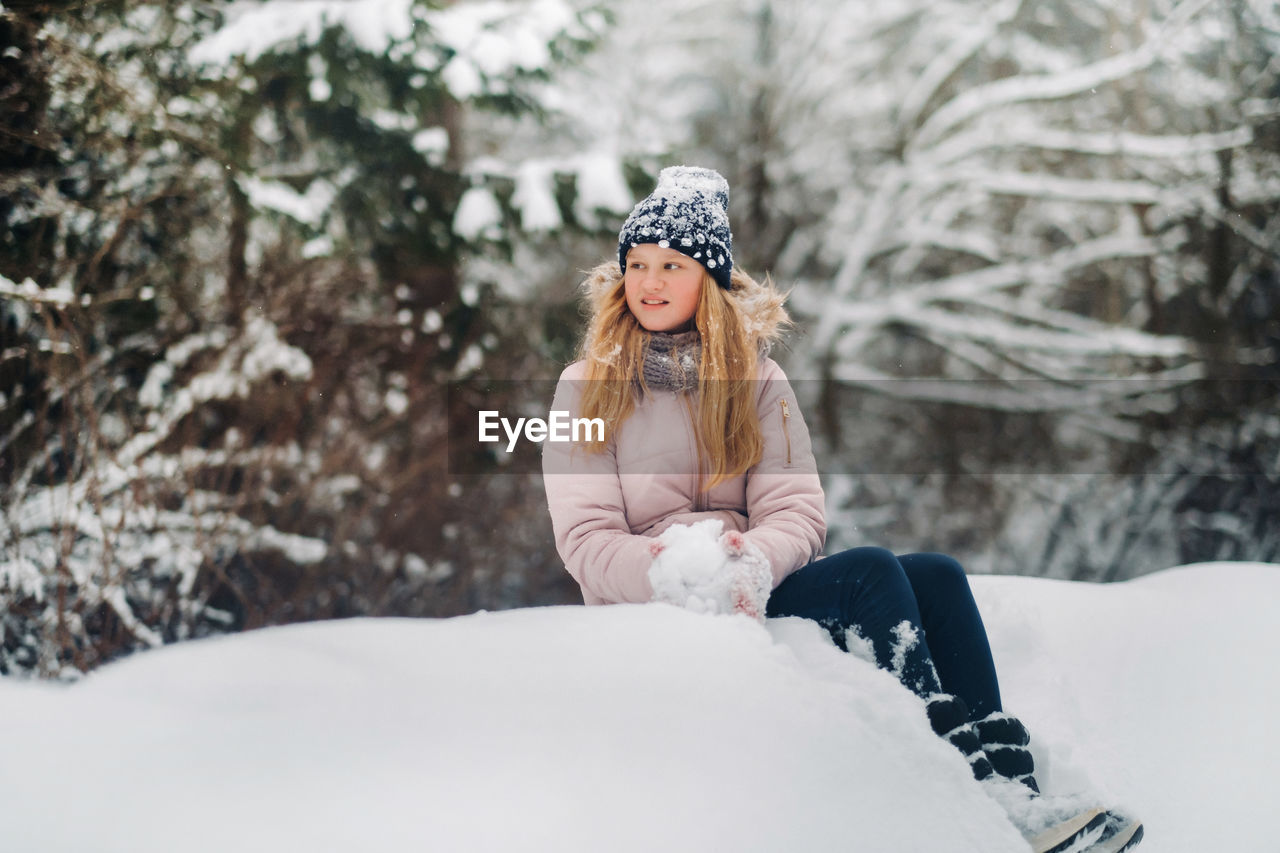 Cute girl with long hair is sitting on a mountain of snow in a hat and making snowballs