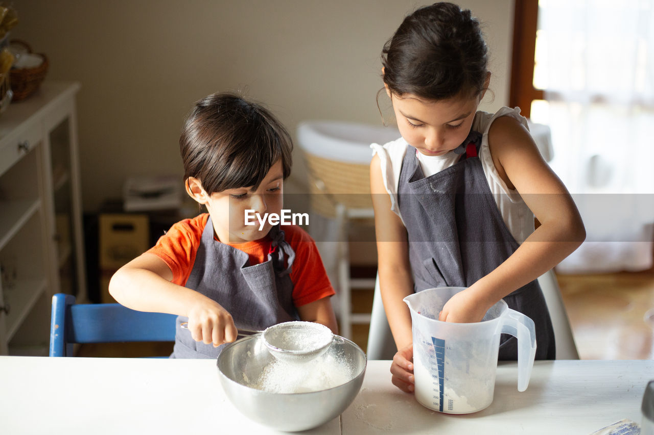 Two children wearing grey aprons sifting flour