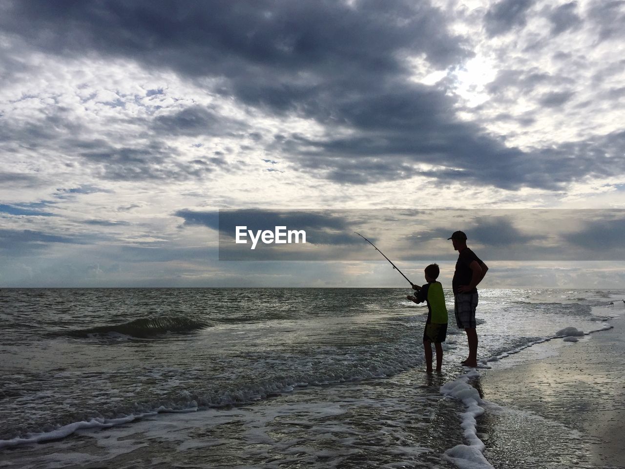 Father and boy fishing on shore at beach against sky