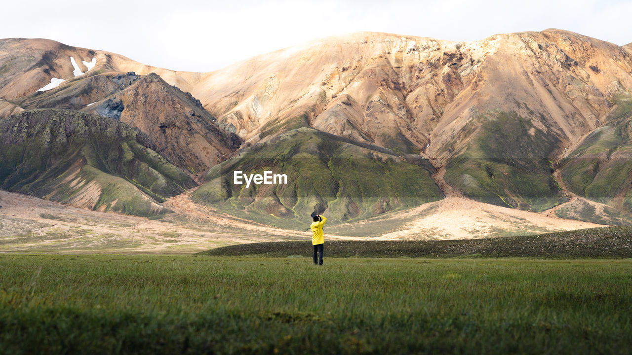 REAR VIEW OF MAN STANDING ON MOUNTAIN AGAINST MOUNTAINS