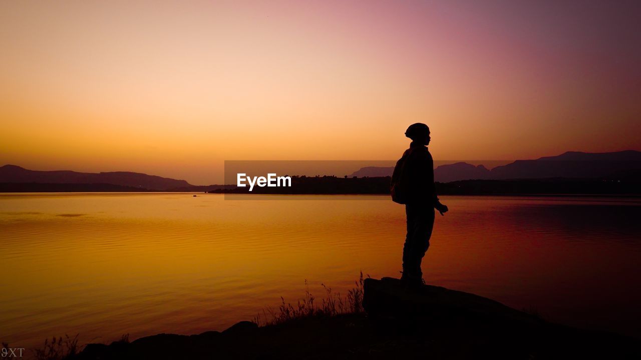 Silhouette man standing on rock by lake against orange sky