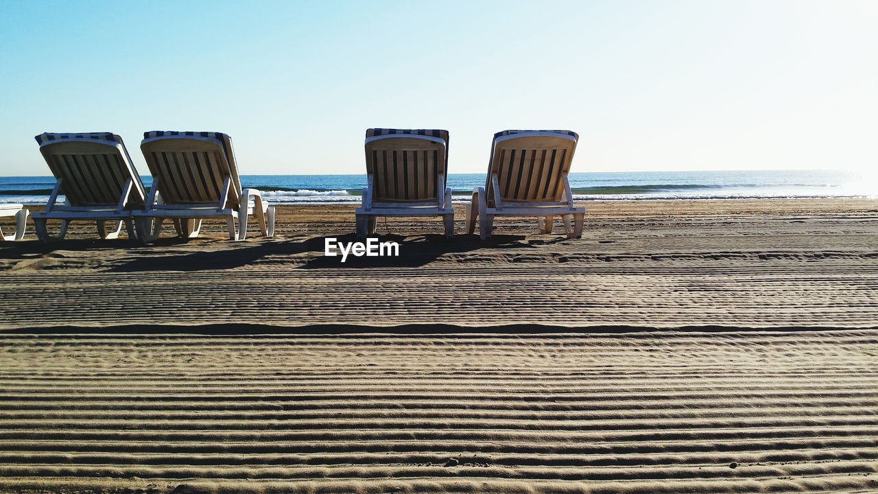 Empty deck chairs on sand at beach against clear sky