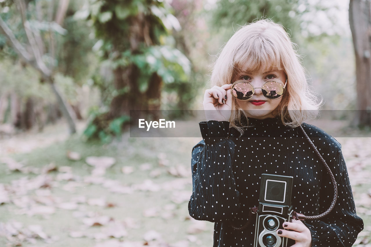 Portrait of young woman wearing sunglasses while holding vintage camera in park