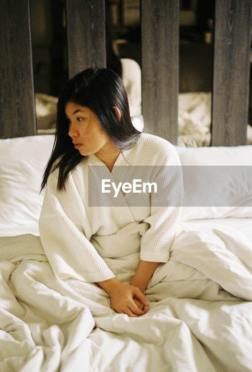 Thoughtful woman wearing white bathrobe while sitting on bed at home