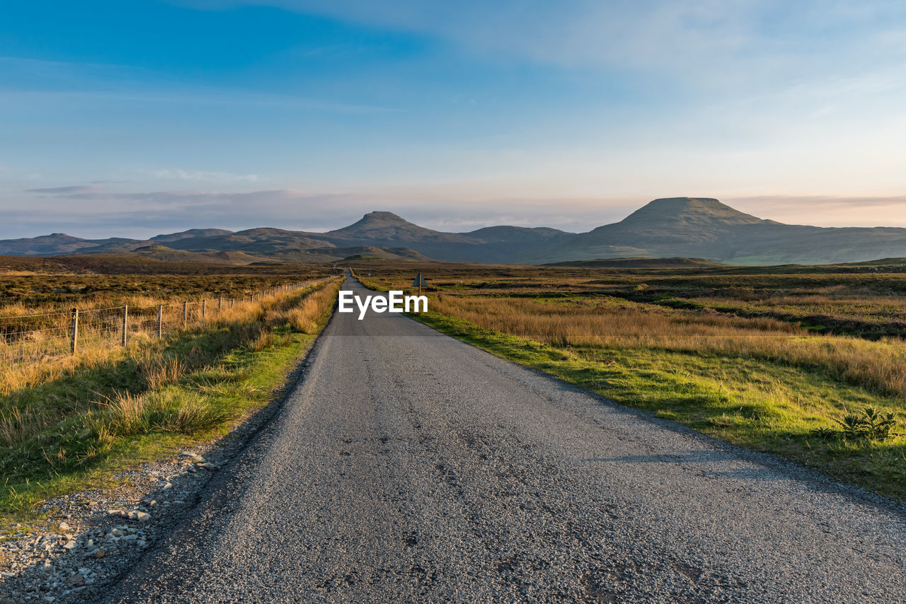 A straight, narrow and empty tarmac road meets at the horizon with the mountains at the isle of sky