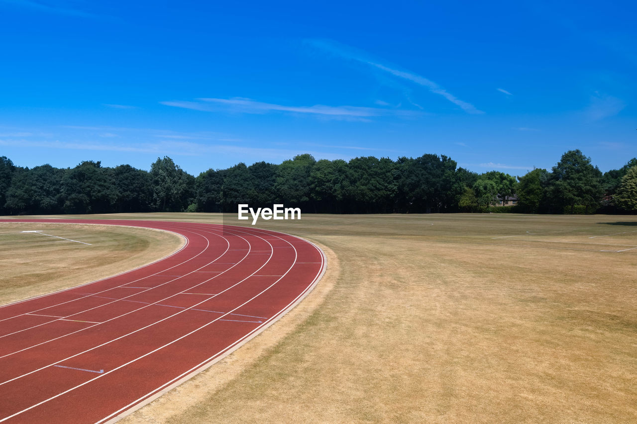 Empty athletics running track amidst field and trees on horizon against blue sky with copy space 