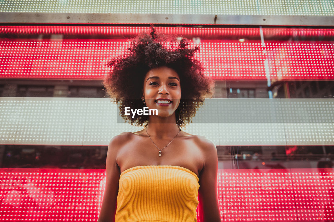 Low angle portrait of happy young woman standing against neon american flag in city