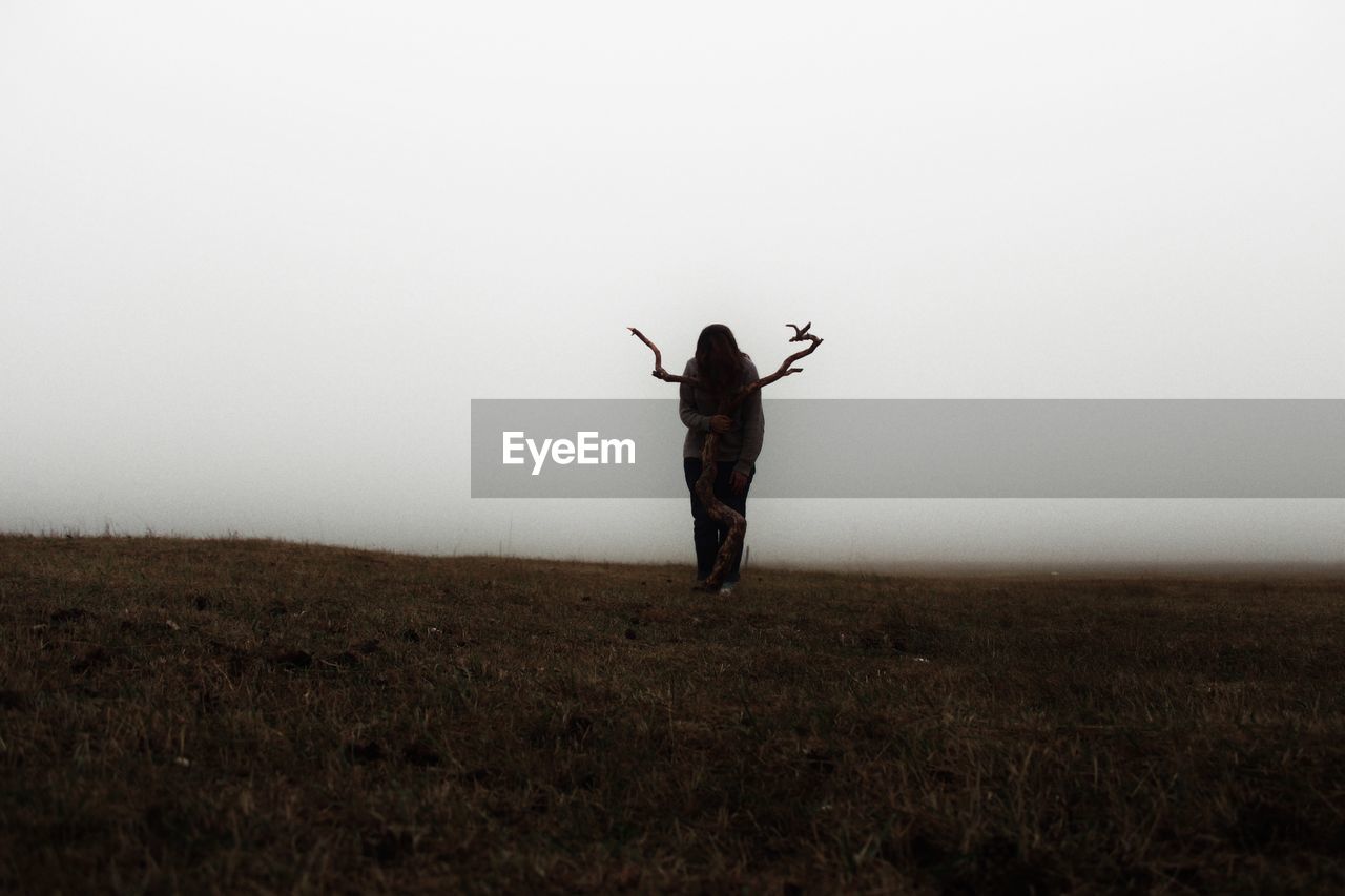 Young woman standing with wood on field against clear sky