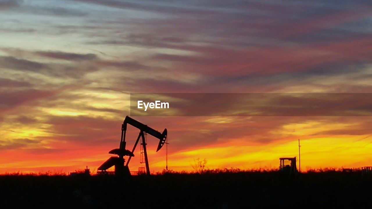 Silhouette machinery on oil field against cloudy sky during sunset