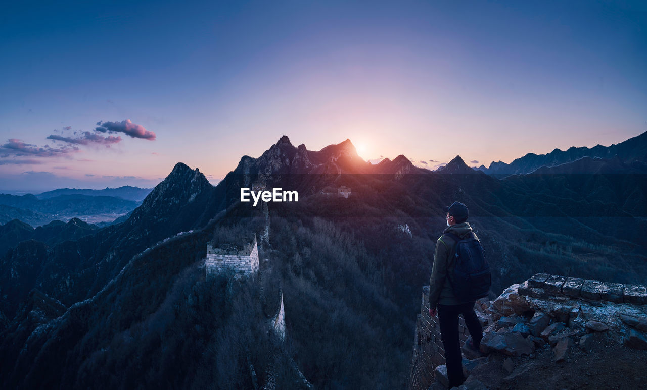 Man with backpack looking at view while standing on cliff against mountain and sky during sunset