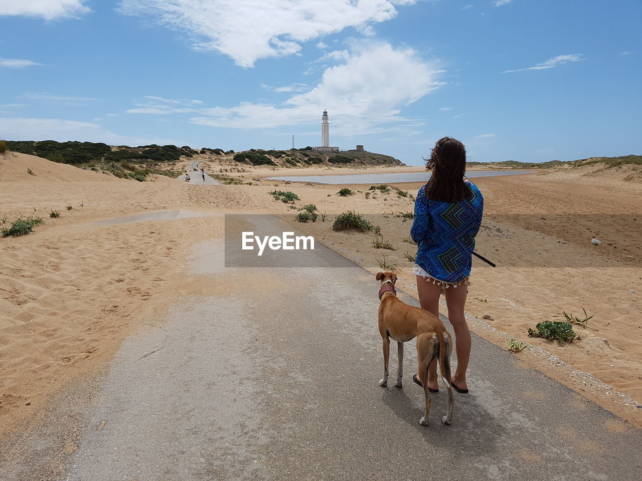 Woman with greyhound standing on road leading towards lighthouse against sky