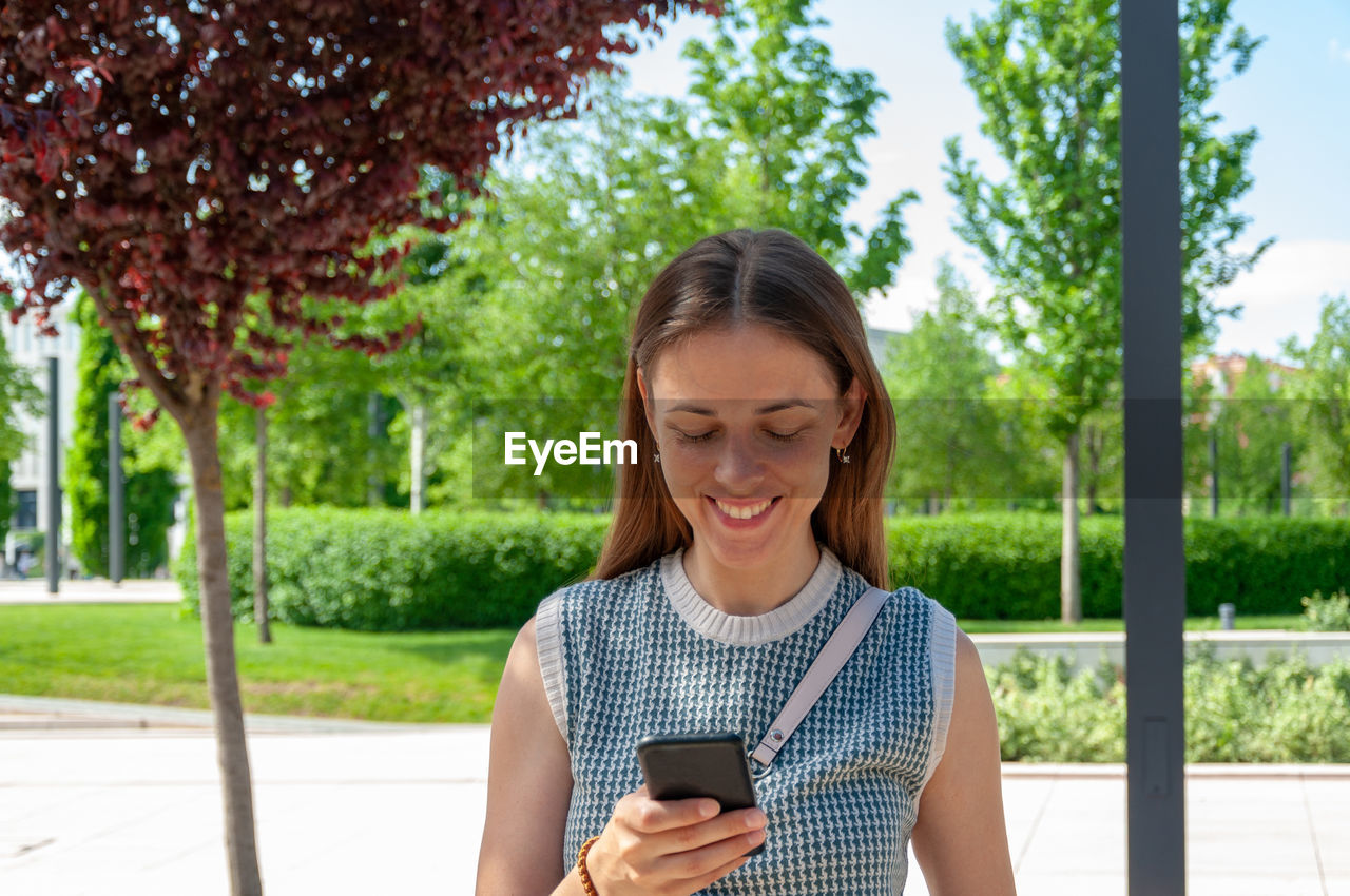 BEAUTIFUL YOUNG WOMAN USING PHONE WHILE STANDING BY PLANTS