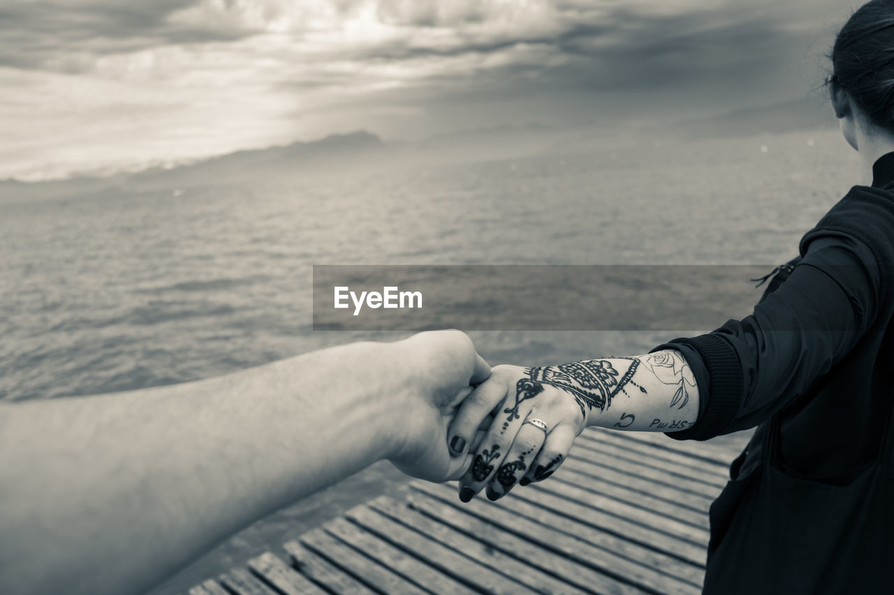 Cropped hand on man holding girlfriends hand while standing on pier against cloudy sky